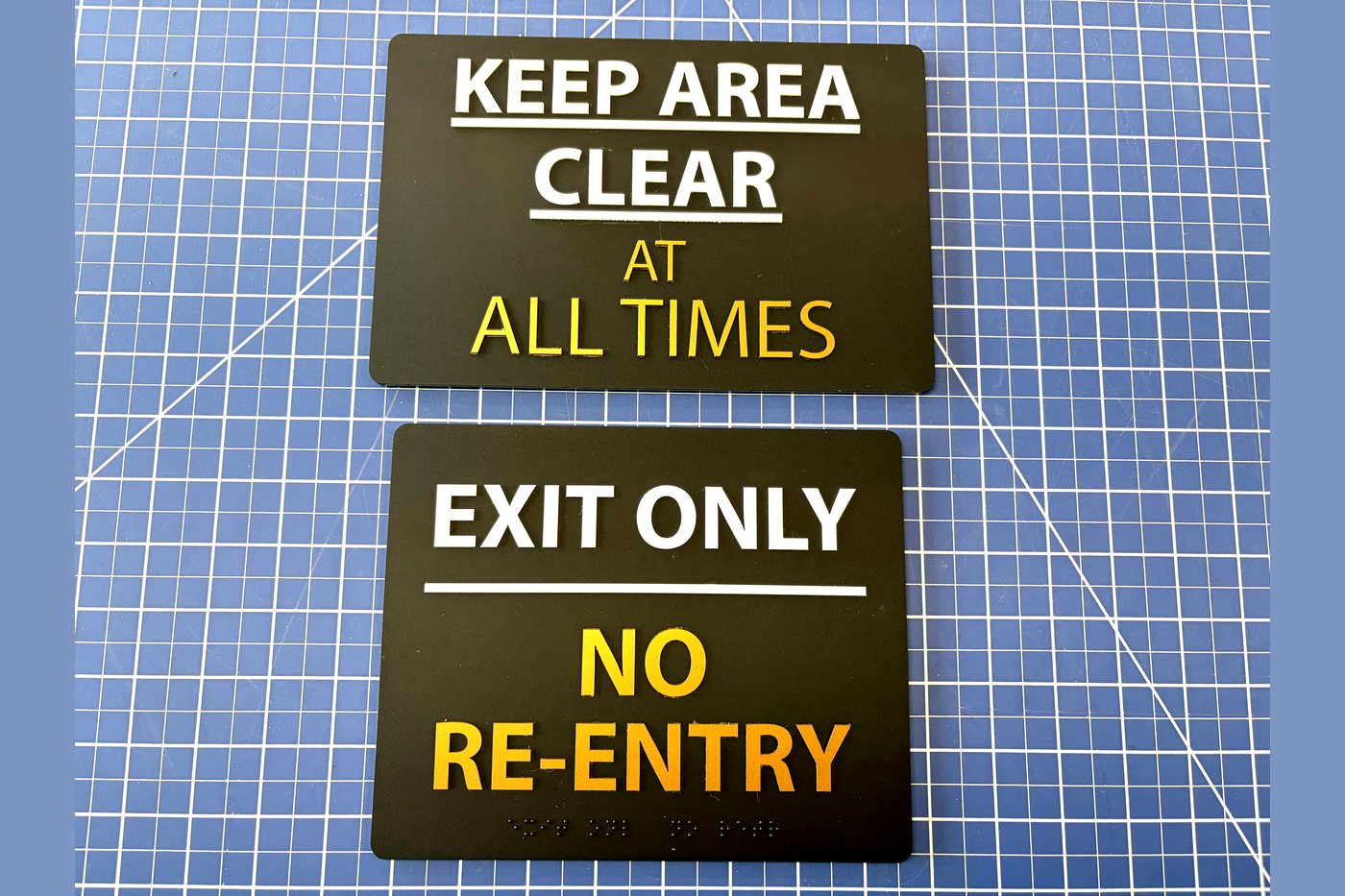 Two black informational signs with raised ivory and orange characters, Keep Area Clear At All Times and Exit Only No Re-Entry